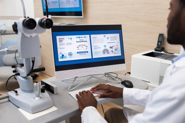 An ophthalmologist wearing a white lab coat is seated at a workstation, working on a computer displaying an eye care consultation interface. The screen shows information about ophthalmology services, including clinic consultation details and eye care solutions. Beside the monitor, there is an ophthalmic examination device, suggesting a medical or optometry setting. The background features a printer, a phone, and a wall-mounted screen displaying similar content